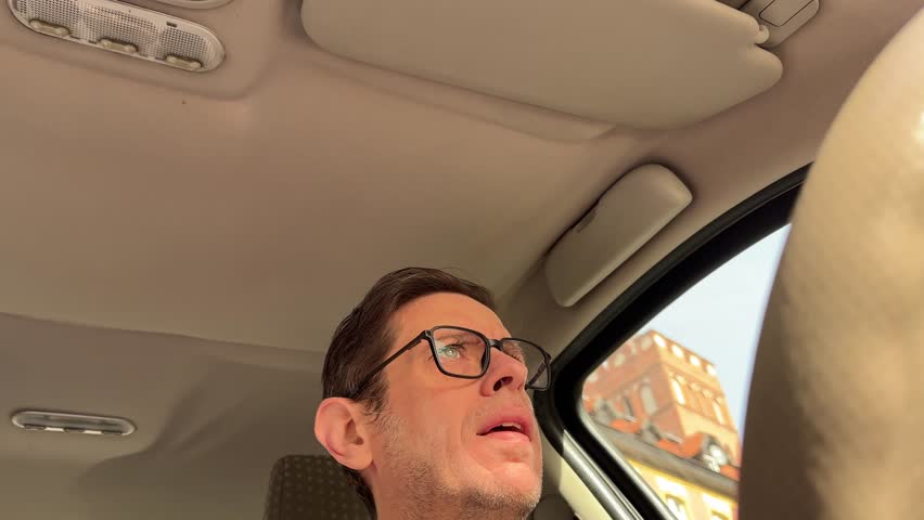 Video footage of a middle aged man driving a car seen from inside the vehicle with natural daylight providing an authentic everyday scene for stock footage.