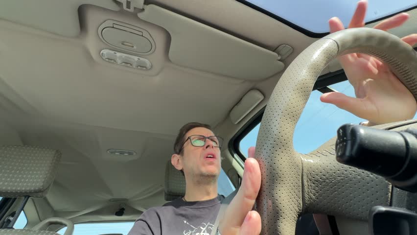 Footage of a middle aged man driving a car and gesturing with hands inside vehicle interior during a daytime trip on the road.
