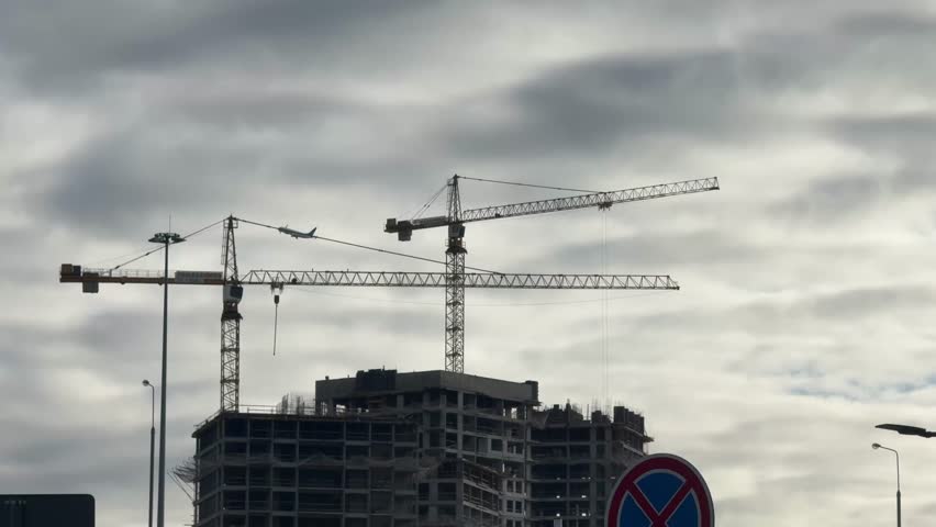 An airplane flying over a modern construction site with two large tower cranes and an unfinished residential building against a cloudy sky. Capturing urban development and real estate growth.