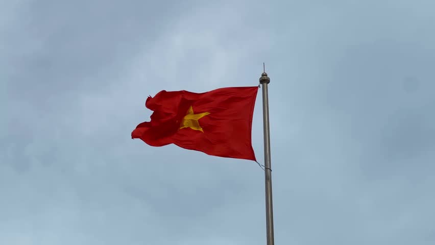 Close up of the Vietnamese national flag waving on a tall flagpole against a cloudy sky, patriotic symbol of Southeast Asia nation.