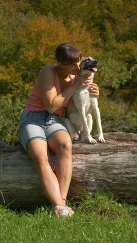 VERTICAL, PORTRAIT: Cheerful young woman hugs her happy mixed breed dog as they sit together on a large fallen log in forest meadow, surrounded by colourful autumn trees and bathed in warm sunlight.