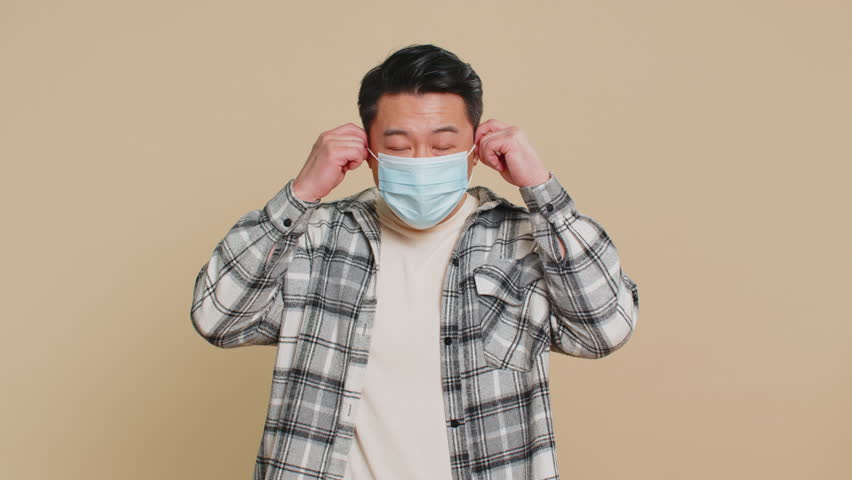 Asian middle-aged man happily removing, taking off medical mask and throws it away. Flu treatment cure. Ending of pandemic quarantine, recover after contagious disease. Chinese guy on beige background
