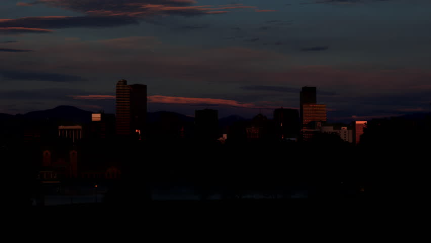 Tight shot of the downtown Denver Colorado skyline illuminated by warm sunrise light, viewed across a calm lake with trees in the foreground. The city buildings gradually brighten as the sun rises, casting golden tones on the architecture while soft clouds drift across the sky. The distant Rocky Mountains provide a layered backdrop, adding depth and regional context to the scene.

The still water reflects portions of the skyline and surrounding landscape, creating a balanced composition with natural and urban elements. The time-lapse sequence emphasizes changing light, shifting shadows, and the transition from early morning to full daylight.

This footage is ideal for projects related to urban environments, travel, business, and lifestyle themes. It works well for establishing shots, corporate videos, tourism promotion, and editorial storytelling. The clean composition also allows for easy integration of text or graphics.

The scene conveys themes of growth, calm beginnings, modern development, and connection between nature and city life. High-resolution footage suitable for professional video production and commercial use.

Concepts include cityscape, skyline, sunrise, morning light, downtown, architecture, urban landscape, mountains, lake, reflection, Colorado, travel, business, and environment.
