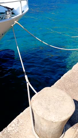 A white boat is secured to a concrete bollard on a dock by thick mooring ropes.The deep,clear turquoise water of the sea gently undulates, creating a high-contrast maritime scene under bright sunlight