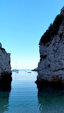 A sailboat anchors outside the narrow entrance of a stunning cove or gorge, framed by towering, layered limestone cliffs. The calm, clear water reflects the blue sky, capturing the secluded beauty