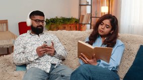 Indian couple man and woman sitting on home sofa, spending calm day together as girl reads book and man enjoys music in headphones. Young family sharing peaceful time side by side in relaxed mood. - Powered by Shutterstock - Get 15% off with code: PIKWIZARD15