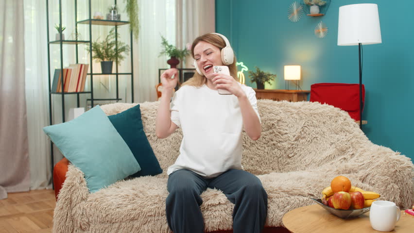 Pregnant middle-aged woman at home dances holding smartphone like microphone sings hit song full of joy. Caucasian girl on sofa moves feet shakes head laughs feels music so happy energy waiting baby.