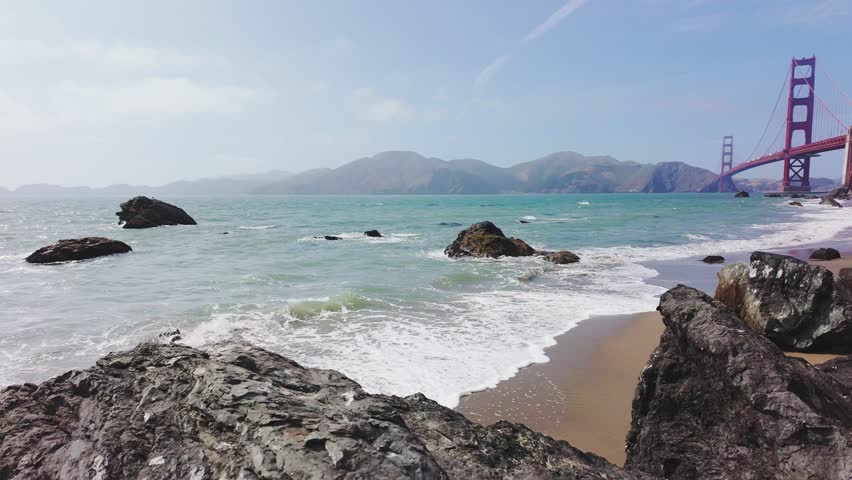 Waves roll onto sandy shore at Baker Beach with Golden Gate Bridge in background, San Francisco, California, USA. Iconic coastal view with ocean, rocks and Marin Headlands