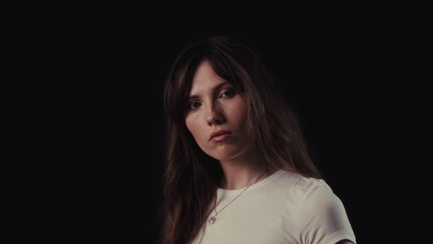 Studio portrait of a beautiful young woman with long brown hair, wearing a white t shirt, adjusting her hair on a dark background