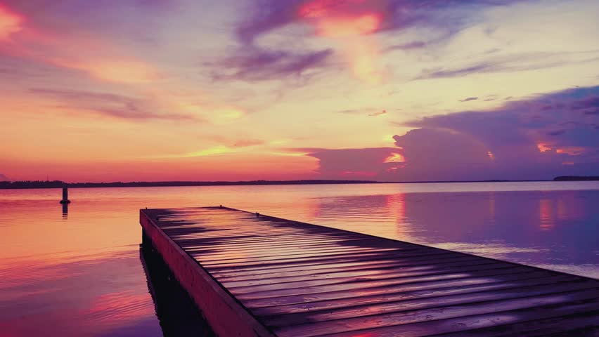 Experience the serene beauty of a sunset over a tranquil lake. A wooden dock extends into the calm waters, reflecting the vibrant colors of the sky. Enjoy this peaceful scene.
