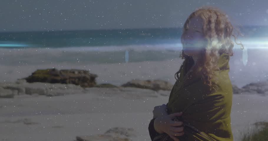 Woman wrapping coat on windy beach, adjusting shawl for warmth while flare enhancing travel scene. Seaside, solitude, breeze, sunlight, contemplative, nature, outdoor