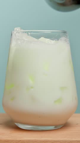 Vertical video close up: matcha stream pours into iced milk in glass on wooden board, distinct layers form, beverage preparation step.