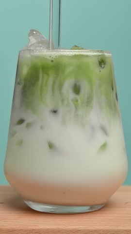 Vertical closeup: barista in glove stirs iced matcha latte with glass straw in tumbler, layers blend to smooth green drink.