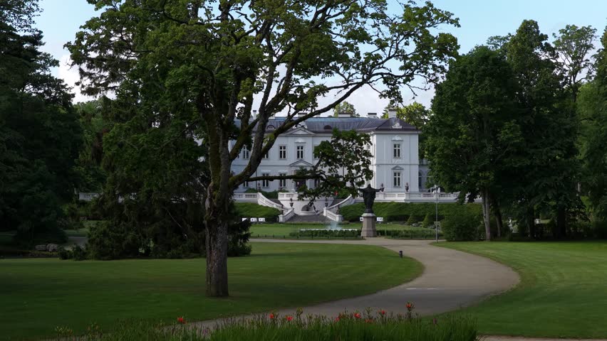 Aerial view of a neoclassical mansion surrounded by lush greenery in Palanga, Lithuania. The estate features manicured gardens, a central fountain, and a statue in the foreground.