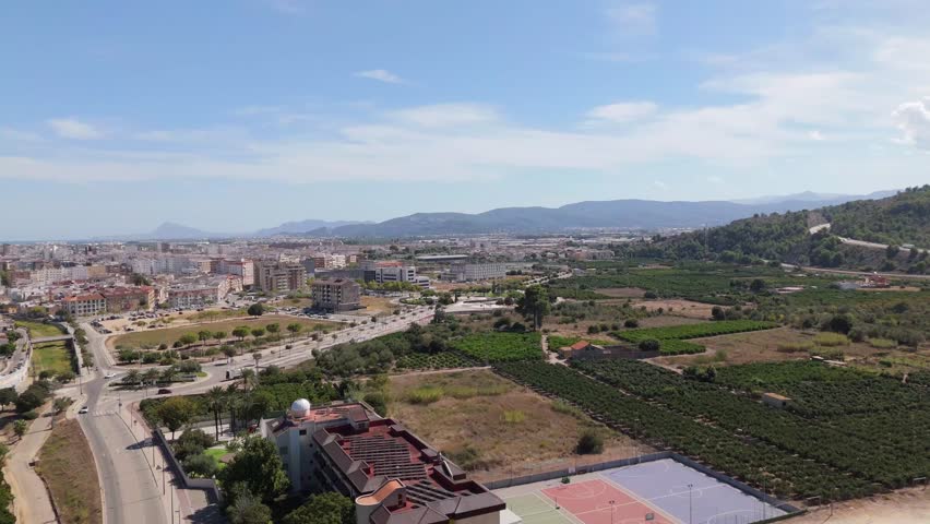 Aerial drone view of Gandia, Spain, showing modern buildings, a skatepark, tree-lined streets, and surrounding mountains in the background.