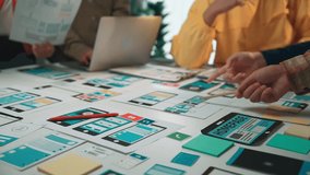 A dynamic team collaborates on mobile app design, engaging in discussion and sharing ideas around colorful wireframe mockups in a bright office environment. SACTR - Powered by Shutterstock - Get 15% off with code: PIKWIZARD15