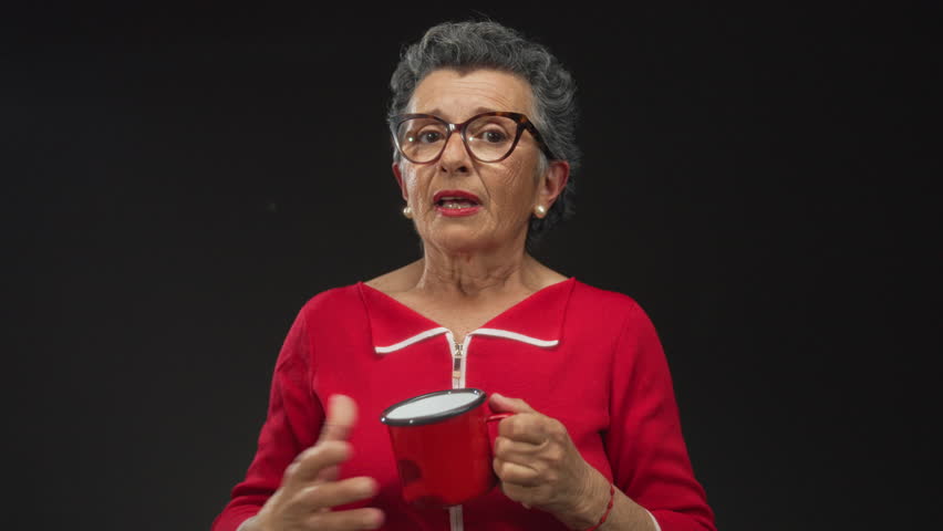 Senior woman with grey hair looks concerned while holding a red coffee cup against a black background, wearing glasses and a red shirt.