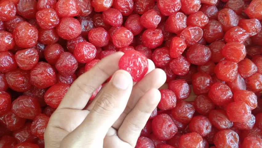Hand holding a bright red cherry above a pile of glossy, wrinkled cherries—vibrant, textured close-up perfect for food, market, or culinary imagery.

