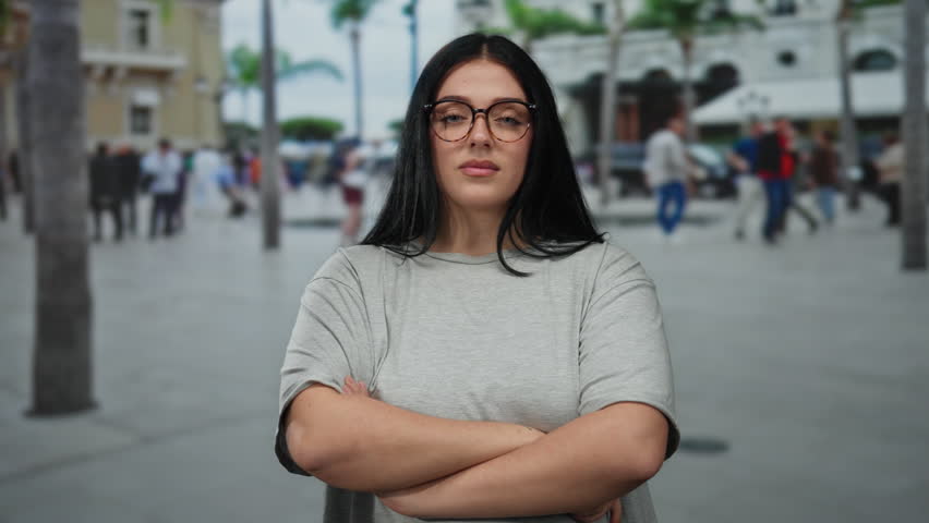 Young woman stands confidently on a busy urban street, showcasing her plus size curvy figure and stylish glasses against a vibrant city backdrop.