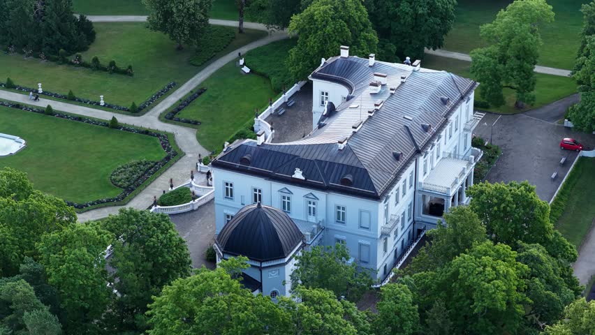 Aerial view of a neoclassical mansion surrounded by lush greenery in Palanga, Lithuania. The estate features manicured gardens, a central fountain, and a statue in the foreground.