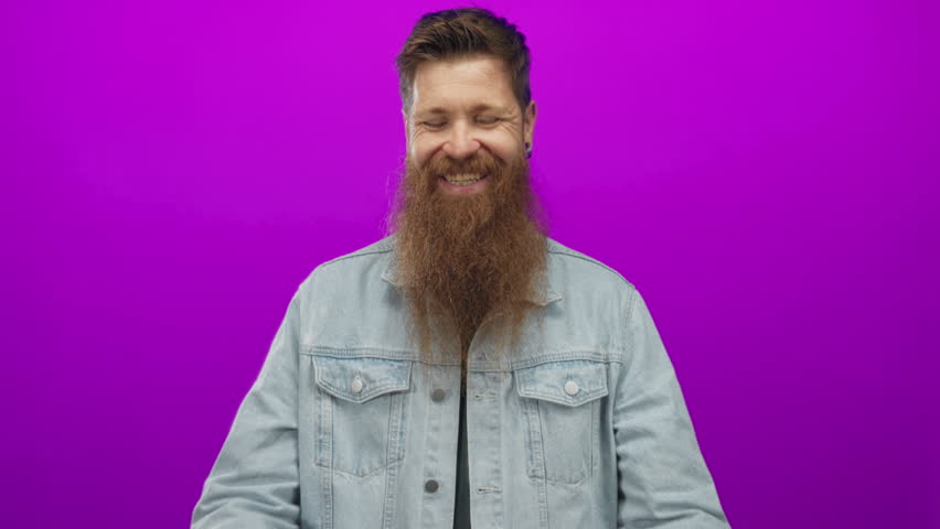 Man clapping hands in studio with a vibrant purple backdrop, wearing denim jacket and full beard; celebration appreciation gratitude joy.