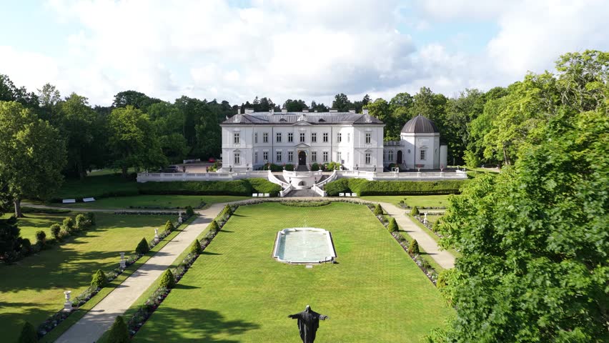 Aerial view of a neoclassical mansion surrounded by lush greenery in Palanga, Lithuania. The estate features manicured gardens, a central fountain, and a statue in the foreground.