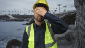Man builder covering eyes with hand by a coastal building near rocky shore wearing hardhat and high vis vest; fatigue. - Powered by Shutterstock - Get 15% off with code: PIKWIZARD15