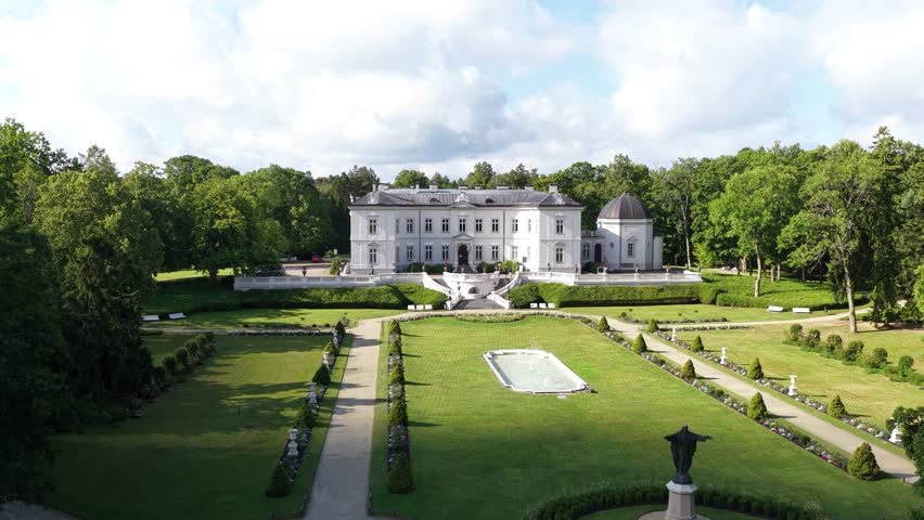 Aerial view of a neoclassical mansion surrounded by lush greenery in Palanga, Lithuania. The estate features manicured gardens, a central fountain, and a statue in the foreground.