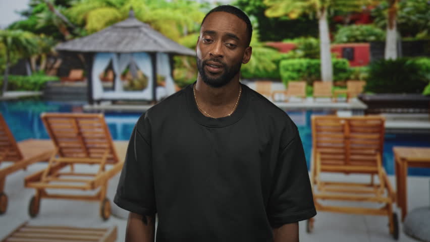 Man clutching chest with hand on sternum at resort pool beside hotel building, standing near lounge chairs; concern.