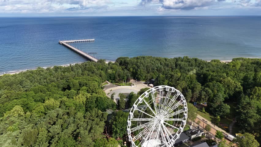 Elevated view of a Ferris wheel rising above lush green trees in Palanga, Lithuania, with a church tower and city buildings visible in the background.