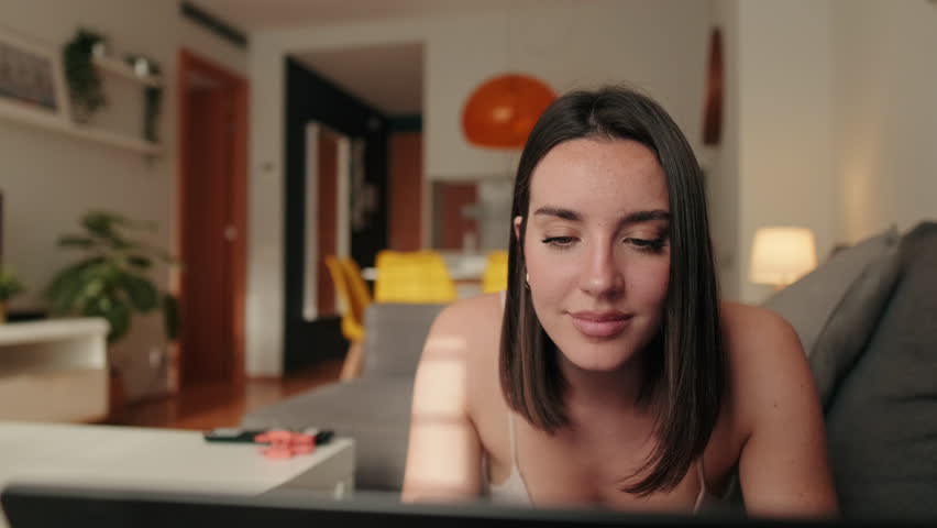 Young woman lying on her couch at home, browsing on laptop computer. Sunlight on her face. Comfortable and focused.