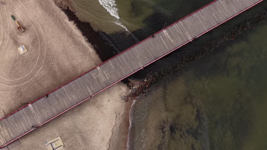 Close-up view of a wooden pier with red and blue railings stretching into the Baltic Sea in Palanga, Lithuania, with people walking along the structure.