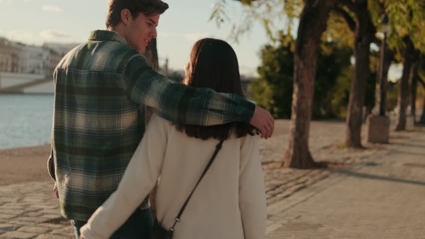 A young couple walks along the Guadalquivir River in Seville, Spain, arm in arm. They kiss and talk as they stroll along the scenic promenade.