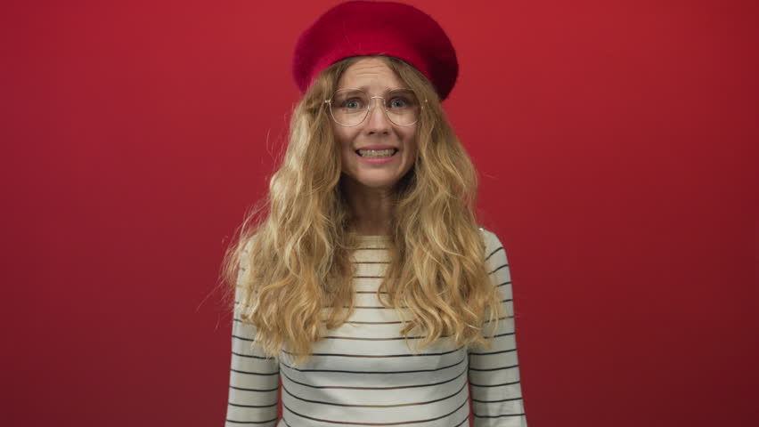 Woman hands covering her eyes in a studio shoot showcasing a blonde young model wearing a beret against a red backdrop with striped attire and glasses.