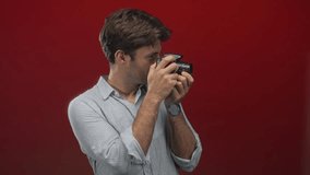Young hispanic man holding a camera and looking through the viewfinder in a studio with a red backdrop and rolled sleeves; concentration creativity. - Powered by Shutterstock - Get 15% off with code: PIKWIZARD15