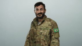 Hispanic man in military uniform points to brazilian flag patch on his sleeve, standing against a grey background with a proud expression and a beard. - Powered by Shutterstock - Get 15% off with code: PIKWIZARD15