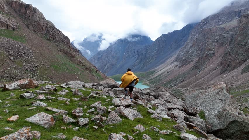 Man wearing a bright yellow rain jacket walks over a rocky foreground, looking down at a stunning turquoise alpine lake nestled between steep mountain slopes and peaks