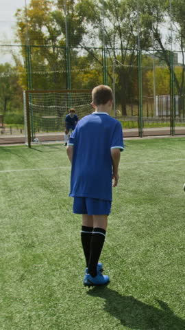 Vertical shot of unrecognizable preteen boy in blue football kit scoring while goalkeeper trying to defend goal