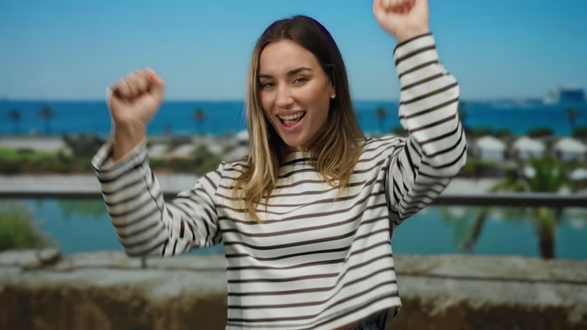 Woman smiling seaside wearing striped shirt standing near ocean boat in lively port outdoor on sunny day with beach scenery and palm trees.