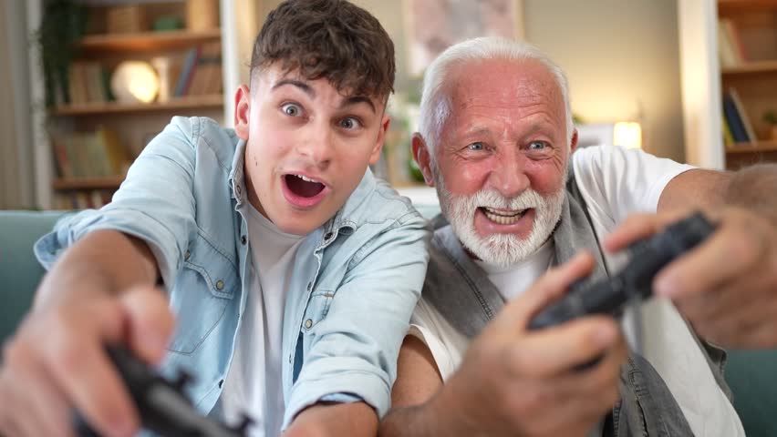 Joyful multi-generational family, an elderly grandfather and a teenage grandson, having fun together, laughing and competing while playing video games with controllers in the living room