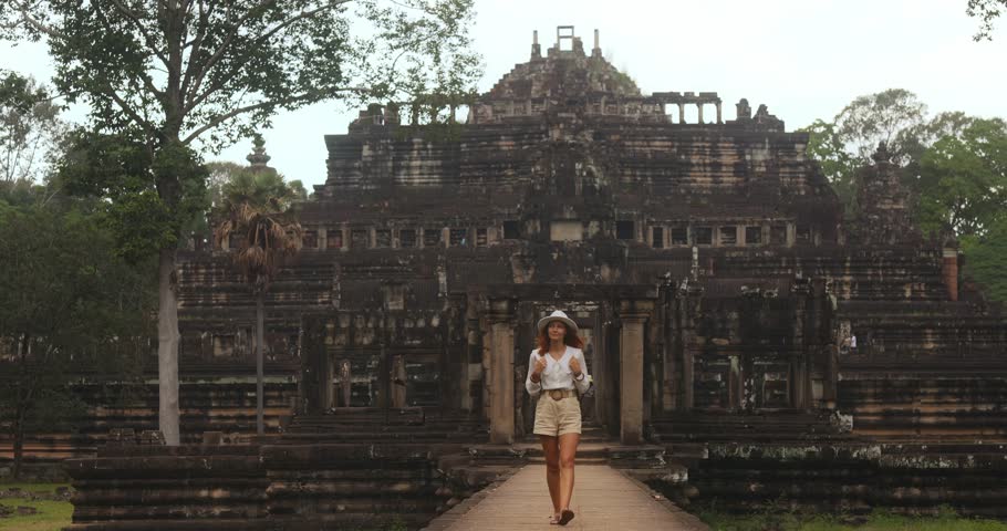 Stylish female traveler in hat and shorts walks from Baphuon Temple toward the camera in Angkor, Cambodia. High quality conceptual stock footage for tourism promos, lifestyle ads, and documentaries.