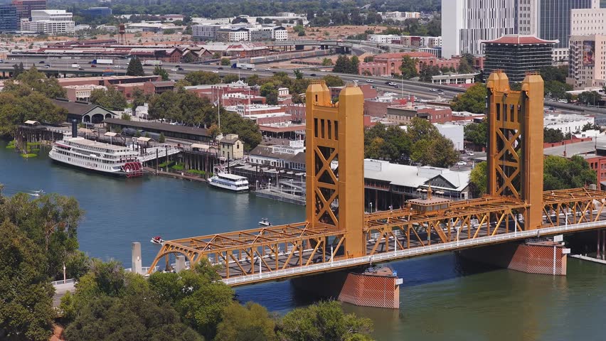 Aerial view of Sacramento, California, featuring the golden Tower Bridge, a boat on the Sacramento River, and a mix of urban and green spaces.
