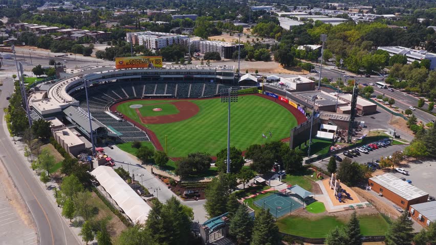 Aerial pan of a baseball stadium in Sacramento, California, showing the field, seating, advertisements, parking lots, and tree lined streets.