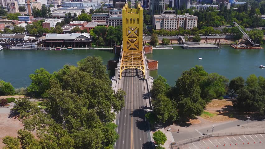 Aerial view of Sacramento, California, featuring the golden Tower Bridge, city skyline, moving boats on the river, and lush greenery on a sunny day.