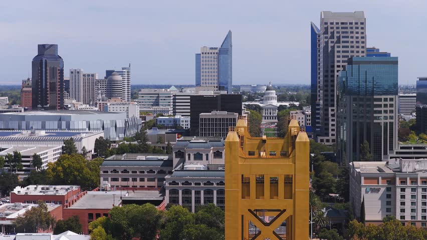 Aerial view of Sacramento, California, featuring the yellow Tower Bridge, State Capitol building, modern skyscrapers, and lush greenery with motion.