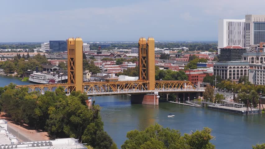 Aerial view of Sacramento, California, showing the golden Tower Bridge, the capitol building, a serene river, and urban architecture with motion.