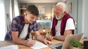 Young grandson assisting his elderly grandfather with filling out documents and using a laptop, providing intergenerational support and guidance with modern technology and financial matters at home - Powered by Shutterstock - Get 15% off with code: PIKWIZARD15