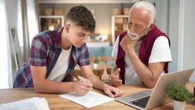 Young man explaining a document to his senior grandfather at the table with a laptop. Grandson helping an elderly relative with financial planning, insurance forms, or paying bills online - Powered by Shutterstock - Get 15% off with code: PIKWIZARD15