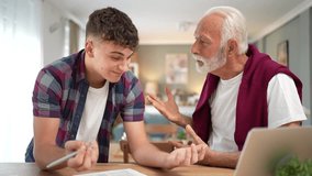 Generational gap showing as an angry grandfather argues with his grandson, who is smiling while writing at a table with a laptop, highlighting a conflict over studying or filling out documents - Powered by Shutterstock - Get 15% off with code: PIKWIZARD15