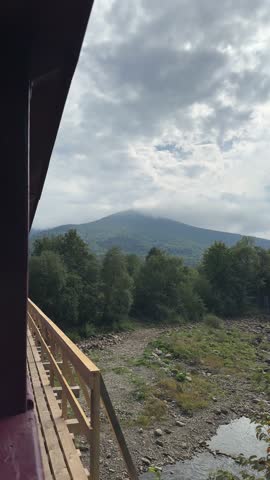 A side view from a train balcony on a wooden bridge offers a misty glimpse of a mountain covered in dense forest with a rocky riverbed below on an overcast day